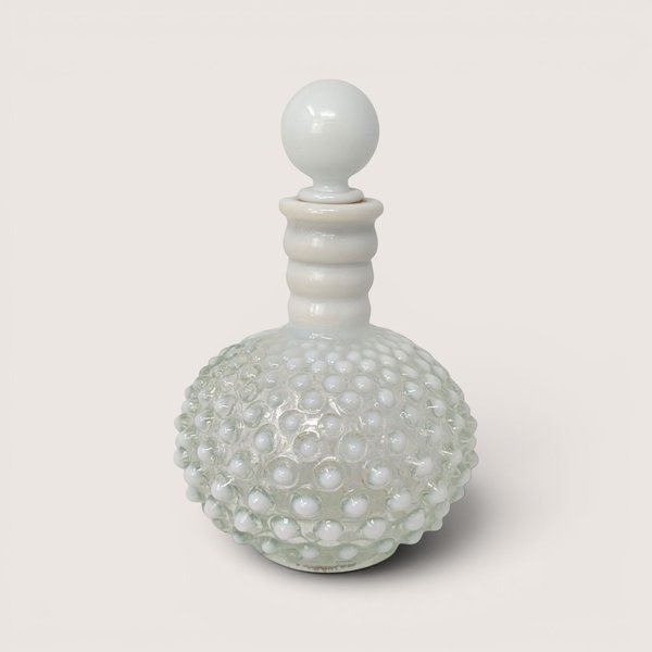 A vintage Fenton white opalescent hobnail cologne bottle with a round ball stopper centered on a light cream background
