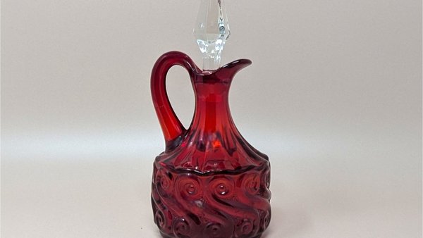A ruby red glass cruet with a bulbous body featuring a raised, swirling scroll pattern. It has a graceful curved handle and a clear, pointed crystal stopper that sits in the narrow neck.