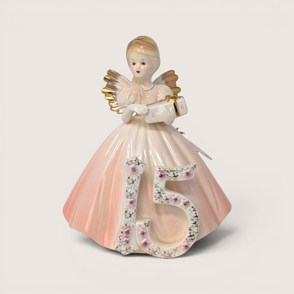 Porcelain birthday angel figurine in a pink dress holding a gift behind a floral number 15 on a solid background.