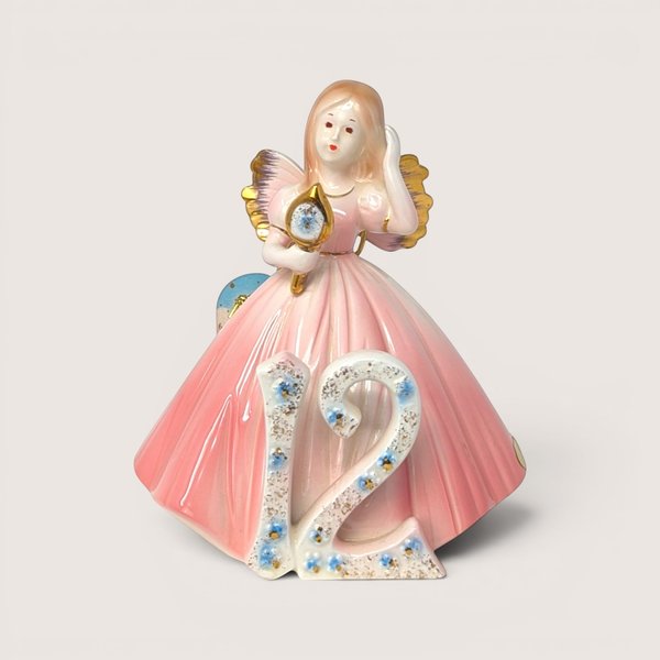 Porcelain birthday angel figurine in a pink gown holding a hand mirror behind a floral number 12 on a solid background.