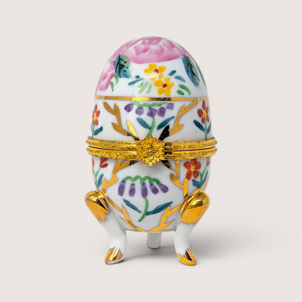 Hand painted floral porcelain egg trinket box with three gold gilded legs and a flower clasp on a solid background.