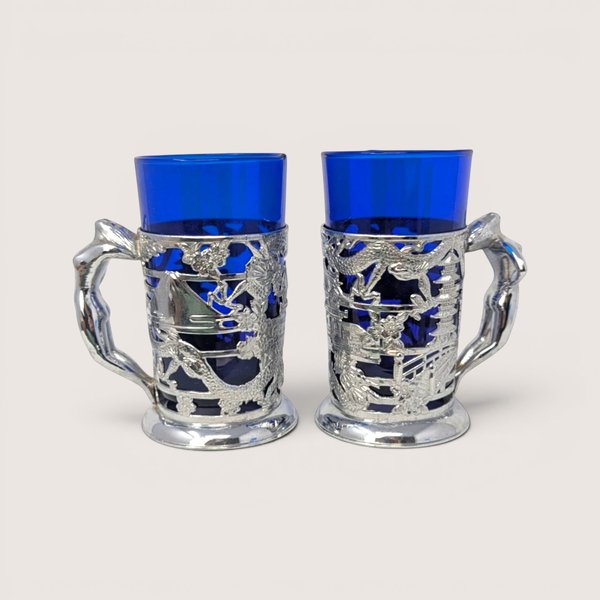Pair of vintage silver plated metal dragon wrap tankards with cobalt blue glass inserts on a solid background.