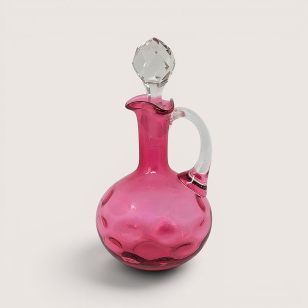 Cranberry glass cruet bottle with a clear applied handle and a faceted clear glass stopper against a neutral background