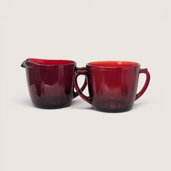 A vintage deep red glass creamer pitcher and matching two handled sugar bowl side by side on a plain background