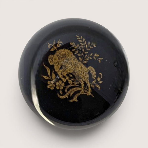 A round dome glass paperweight featuring a detailed gold ram motif over a glossy black lacquer background