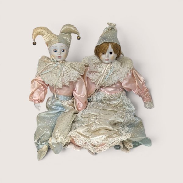 Pair of vintage porcelain Harlequin and Pierrot dolls in pastel pink, blue, and cream satin costumes with lace collars.