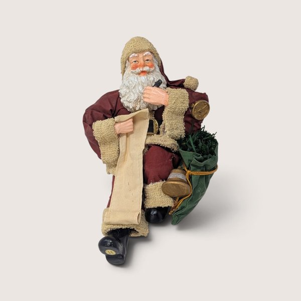 Vintage holiday figurine of Santa Claus sitting down, checking a long list with a pen and holding a toy bag.