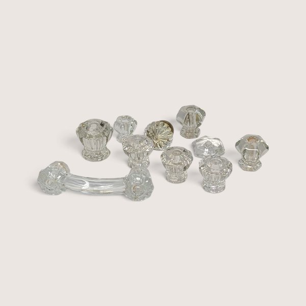 A collection of 10 assorted vintage clear glass cabinet knobs and one curved drawer pull laid out on a neutral background.