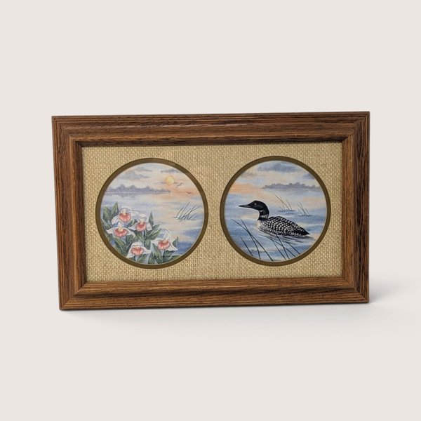 A vintage framed print by D. Smith featuring two circular windows set in a textured fabric mat: one showing white lady's slipper wildflowers and the other a loon swimming on a calm lake, enclosed in a wooden frame.