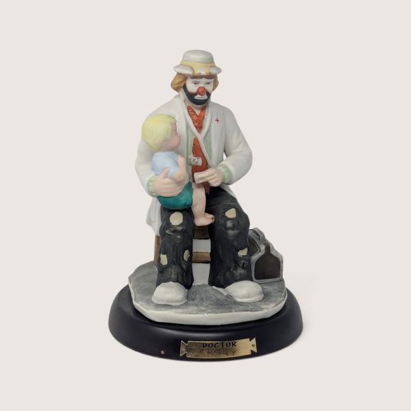 A vintage Flambro Emmett Kelly Jr. porcelain doctor figurine featuring a clown holding a child on a wooden base.
