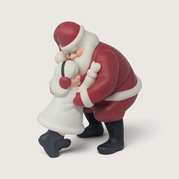 A vintage Department 56 Winter Silhouette ceramic figurine of Santa Claus in a red suit leaning down to embrace a child in a white coat with red earmuffs.