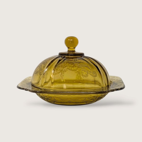 A vintage yellow glass covered butter dish featuring an embossed scroll pattern and domed lid on a neutral background.