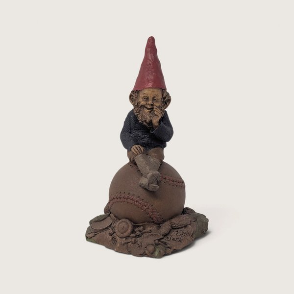 A vintage signed Tom Clark Homer gnome figurine sitting atop a leather baseball on a textured leaf and acorn base against a neutral background.