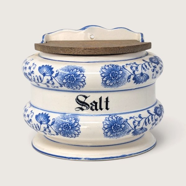 A vintage blue and white ceramic wall-hanging salt bin with a weathered wooden lid and Blue Onion patterns against a neutral background.