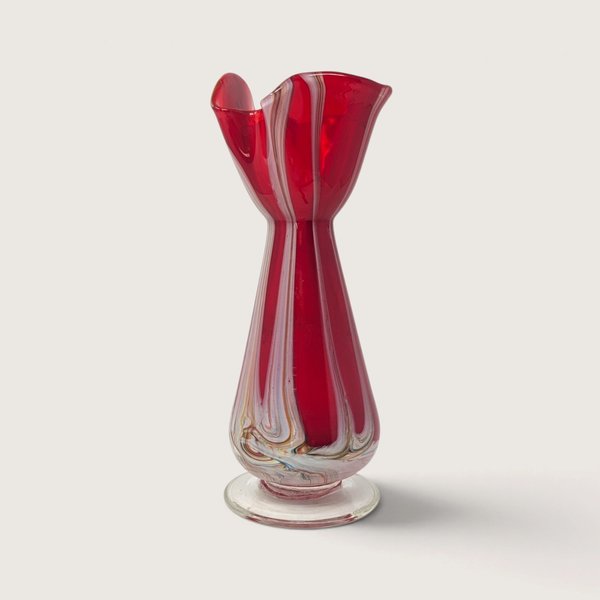A vintage red art glass bud vase featuring white and amber swirl patterns, a flared footed base, and an organic ruffled rim against a neutral background.