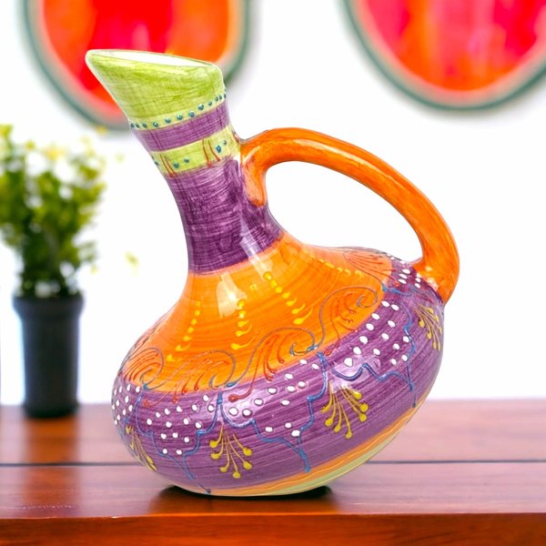 A 9-inch tall handmade ceramic pitcher with a unique slanted or tilted shape, featuring vibrant hand-painted stripes and dot patterns in purple, orange, green, and blue.
