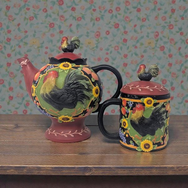 Ceramic decorative teapot and matching lidded mug featuring a red rooster motif and black scrolled borders. The teapot has a curved black handle and spout, while the mug includes a fitted lid.