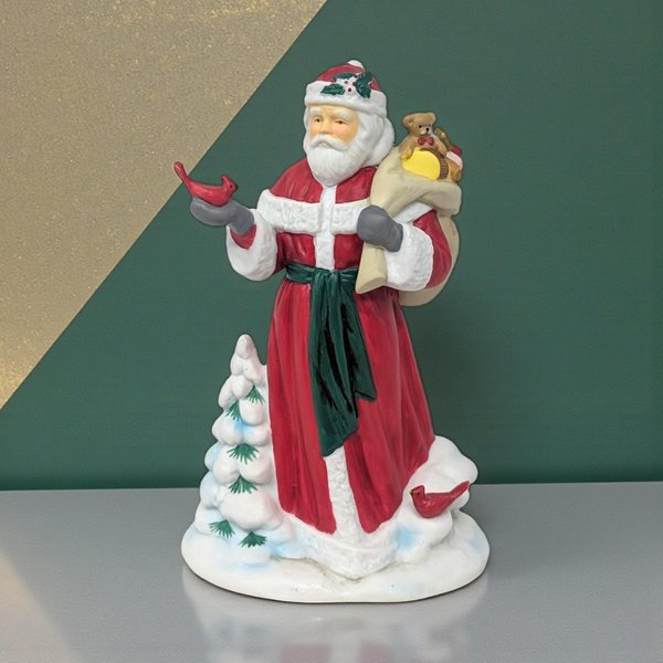Matte porcelain Santa figure in a long red robe with green sash. He holds a red cardinal and a toy sack with a teddy bear. A snow-covered evergreen and second cardinal sit at his base.