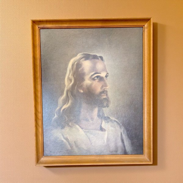 Framed lithograph of the Head of Christ. The portrait features a soft, ethereal glow on the subject's face and long, wavy hair, rendered in muted earth tones and cool blues. The silhouette is set within a simple, honey-toned wooden frame.