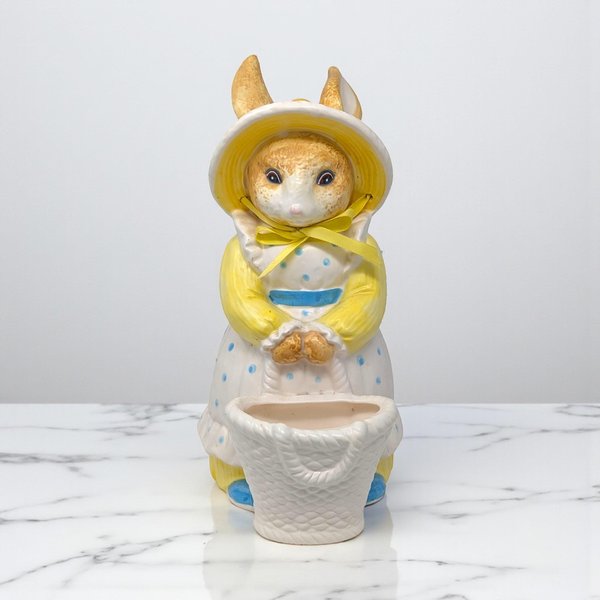 Ceramic bunny figurine wearing a yellow bonnet and a white polka-dot dress with a blue sash. The rabbit holds a small woven-texture basket at its feet. A yellow ribbon is tied in a bow around its neck.