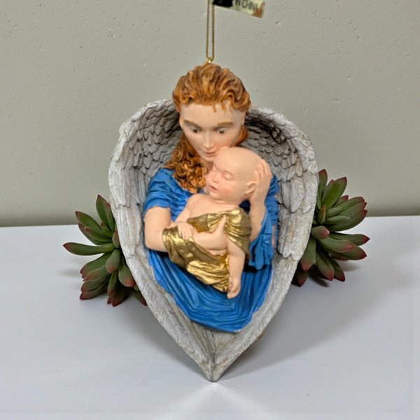 A sculpted resin ornament featuring a red-haired angel in a vibrant blue robe cradling a baby in gold swaddling. The figures are framed by large, textured white wings that form a heart shape at the base.