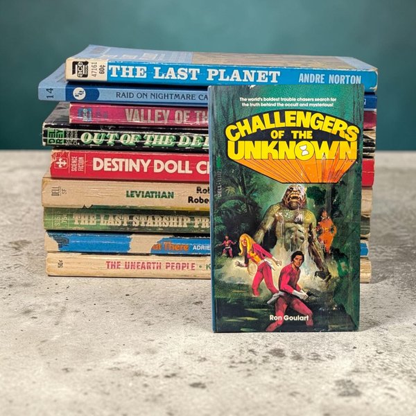 A stack of ten vintage science fiction paperbacks featuring colorful, worn spines. A single book titled "Challengers of the Unknown" by Ron Goulart is leaned against the front of the stack, showcasing dramatic cover art of a giant green monster pursuing two figures in red and pink jumpsuits through a jungle.