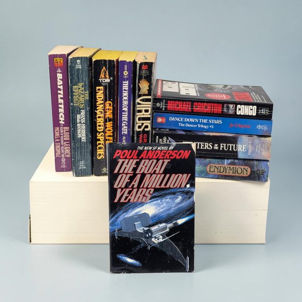 A collection of mass-market science fiction paperbacks is arranged on a white platform. A single book titled The Boat of a Million Years stands in the foreground, featuring a silver spacecraft against a dark blue and black galactic void. The surrounding books display a variety of colorful spines in shades of purple, yellow, and blue, showing visible shelf wear and creased edges along their paper bindings.