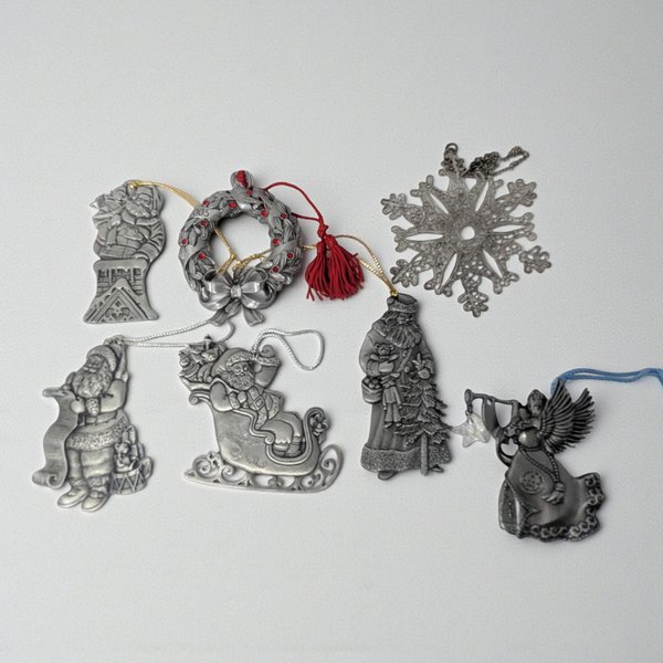 A collection of seven vintage metal ornaments. The set includes detailed pewter figures of Santa in various poses, an angel with a horn, a red-accented wreath with a tassel, and a delicate silver-plated snowflake with an openwork design.