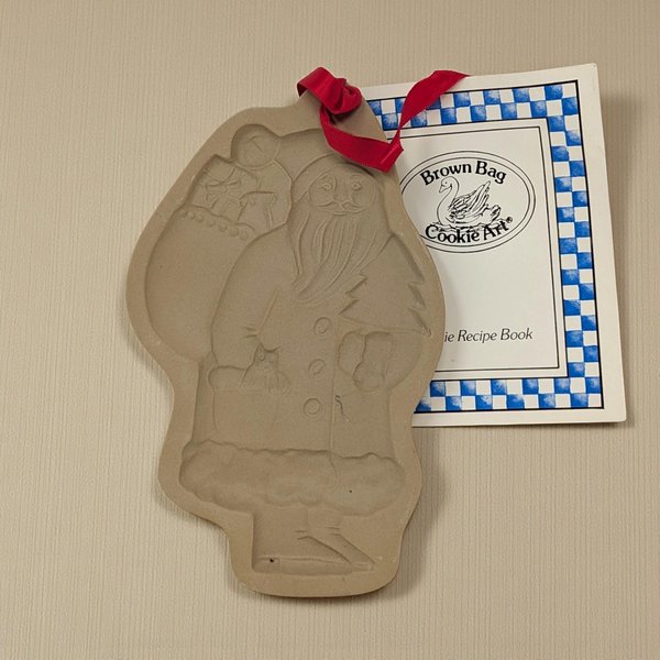 A rectangular stoneware cookie mold featuring a deeply embossed design of a traditional, full-length Santa Claus. He is depicted in a long robe, carrying a sack of toys over his shoulder and a small evergreen tree in his hand, rendered in a warm tan clay.