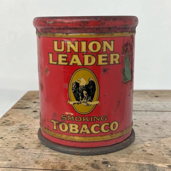 A cylindrical red metal tin with a yellow eagle emblem in the center. Large white block letters read UNION LEADER. A black banner at the top displays SMOKING TOBACCO in yellow font.