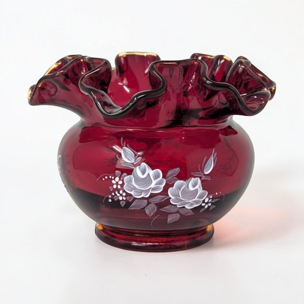 A small, squat glass vase in deep ruby red with a wide, ruffled rim. Delicate white and blue flowers with green leaves are hand-painted on the front. The surface is glossy and smooth with a crimped top edge.