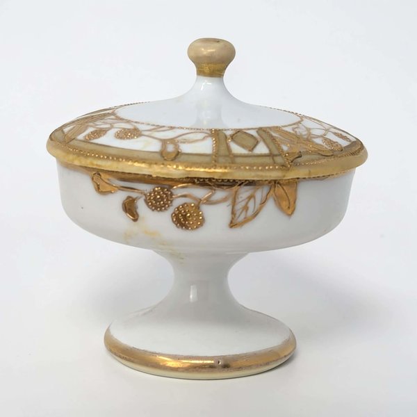 A small, round porcelain lidded box with a creamy white finish. Intricate gold floral patterns and scrolling vine motifs cover the lid and body. The gilding has a slightly raised, textured feel against the smooth porcelain.