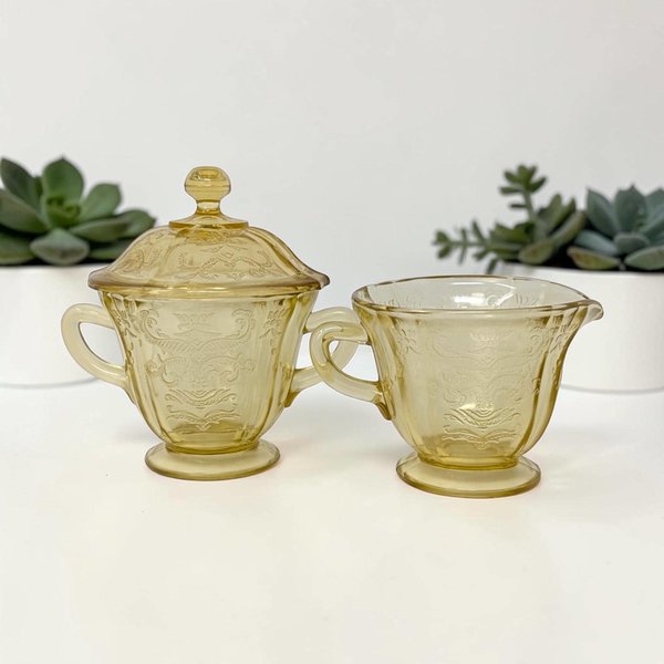 A lidded sugar bowl and matching creamer in warm amber glass. Both pieces feature an intricate, molded lacy scroll and floral pattern. The sugar bowl has a decorative pointed finial on the lid and small handles on either side.
