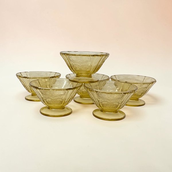 A set of six footed amber glass dessert cups. Each bowl features a flared rim with a lacy, molded scroll and floral pattern. The stems are short and sturdy, leading down to a wide, flat circular base.