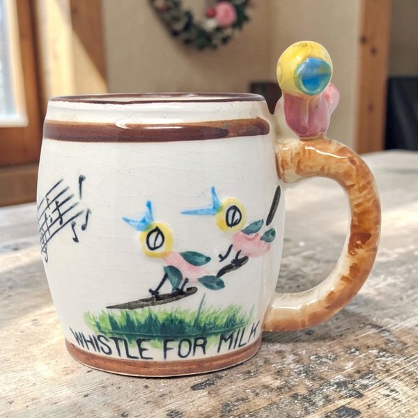 Cream ceramic mug featuring hand-painted whimsical birds on a branch and musical notes. A small sculpted bird sits atop a textured brown handle. Fine crazing visible across the glazed surface with a hand-lettered base.