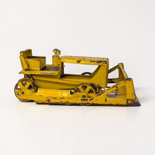 A heavy-duty yellow pressed steel bulldozer with a large front blade and visible surface oxidation. The silhouette is boxy and industrial, featuring a textured open cab and thick black rubber or plastic tracks.