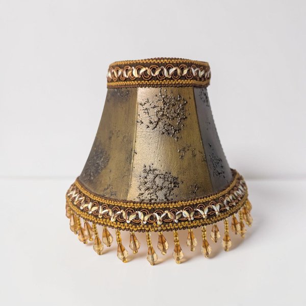 A small, hexagonal clip-on lampshade with a weathered bronze-gold metallic finish and textured distressing. The top and bottom are trimmed with ornate braided cord, featuring amber-colored crystal teardrop beads dangling from the lower rim.