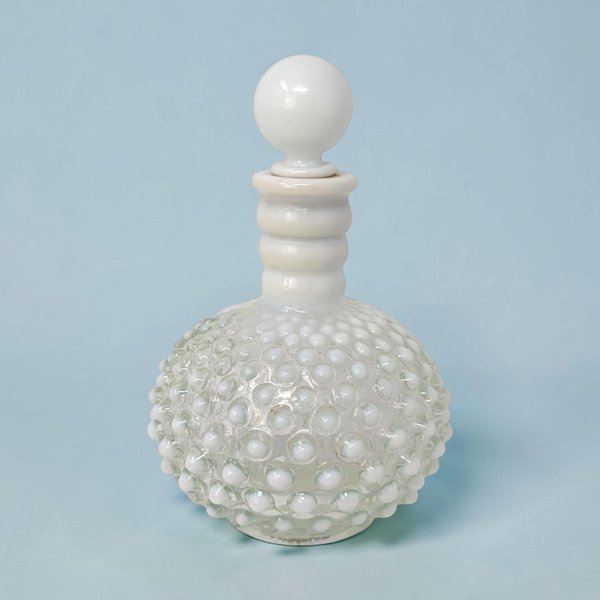 A vintage Fenton white milk glass cologne bottle with a spherical hobnail texture. The bottle features a ribbed neck and a matching smooth round glass stopper on a bulbous silhouette against a light blue background.