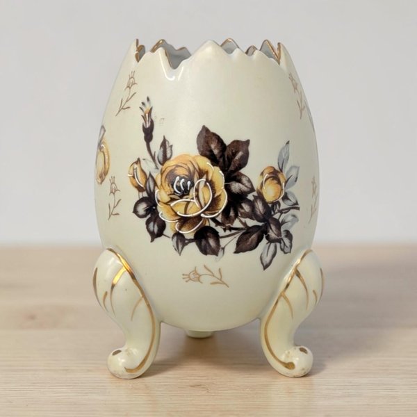 A matte cream porcelain egg-shaped vase with a jagged, broken-egg rim. It features a transfer-ware yellow rose with dark brown leaves and gold-gilded accents on three ornate cabriole feet.