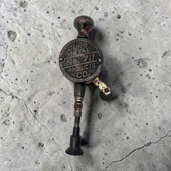 Close-up of a circular vintage metal tool marked Duro Metal Products Co. No. 711 with a dark, weathered patina. A rounded wooden handle sits at the top, and a crank arm with a black knob extends from the side of the industrial-style casing.
