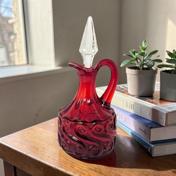 A ruby red glass cruet with a bulbous body featuring a raised, swirling scroll pattern. It has a graceful curved handle and a clear, pointed crystal stopper that sits in the narrow neck.