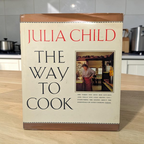 A large, thick hardcover cookbook with a colorful dust jacket featuring photos of prepared food. The texture is a smooth glossy paper over a heavy bound volume with a tall, wide rectangular silhouette.