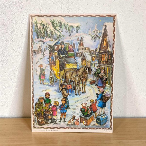 A traditional paper advent calendar featuring a vibrant lithograph of a snow-covered German village at twilight. The rectangular silhouette is accented with delicate iridescent glitter. The numbering on the small perforated doors is clear and crisp.