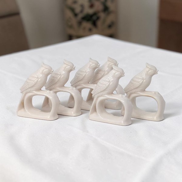 A set of eight matching white porcelain napkin rings. Each ring features a sculpted bird with detailed feather textures, perched on a smooth circular base. The entire piece is finished in a uniform, high-gloss white glaze.