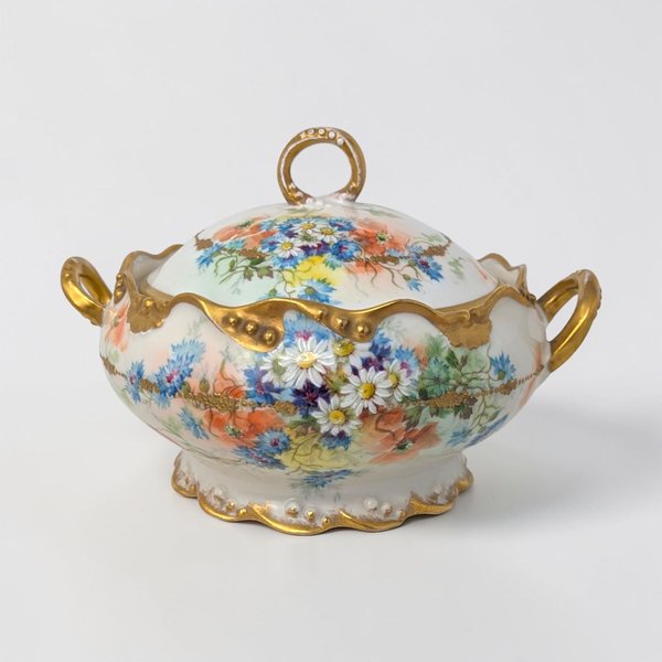 A small, oval white porcelain tureen with a matching lid. It features delicate pink and yellow rose sprays and ornate gold-gilded handles and finial. The elegant silhouette is scalloped along the base and rim.