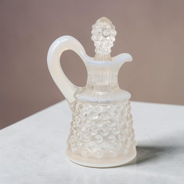 A 4-inch miniature Fenton art glass cruet in white opalescent hobnail. The clear glass base transitions to milky white studs on a flared silhouette, featuring an elegant pouring spout, a curved handle, and a matching hobnail stopper.