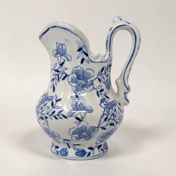A 6-inch ceramic pitcher with a bulbous silhouette and a smooth glazed texture. It features hand-painted blue floral vine patterns on a white background with a scalloped rim and an ornate curved handle.