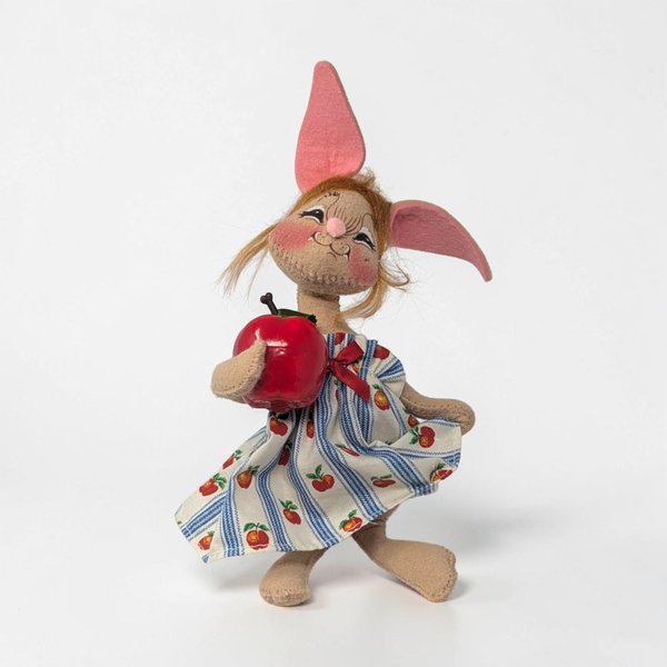 Tan felt bunny figure with a winking face and rosy cheeks. She wears a blue and white striped dress with a red apple print. She holds a large glossy red apple in one hand. Her tall ears are lined in pink felt with blonde mohair tufts.