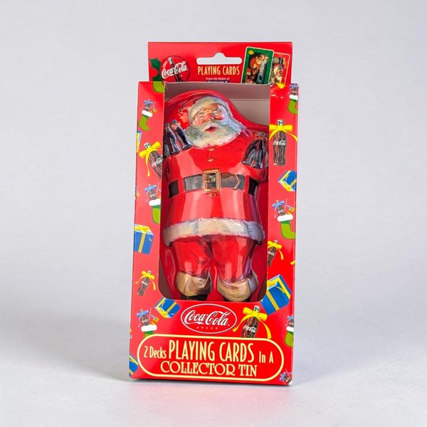 A vertical red metal collector tin featuring a classic illustration of a rosy-cheeked Santa Claus in a red suit holding two bottles of Coca-Cola. The tin is housed in a bright red cardboard display box decorated with colorful gift and stocking motifs.