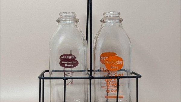 Two vintage clear glass milk bottles with red and orange Rochester Dairy logos sitting inside a black metal wire carrying crate with a wooden handle.